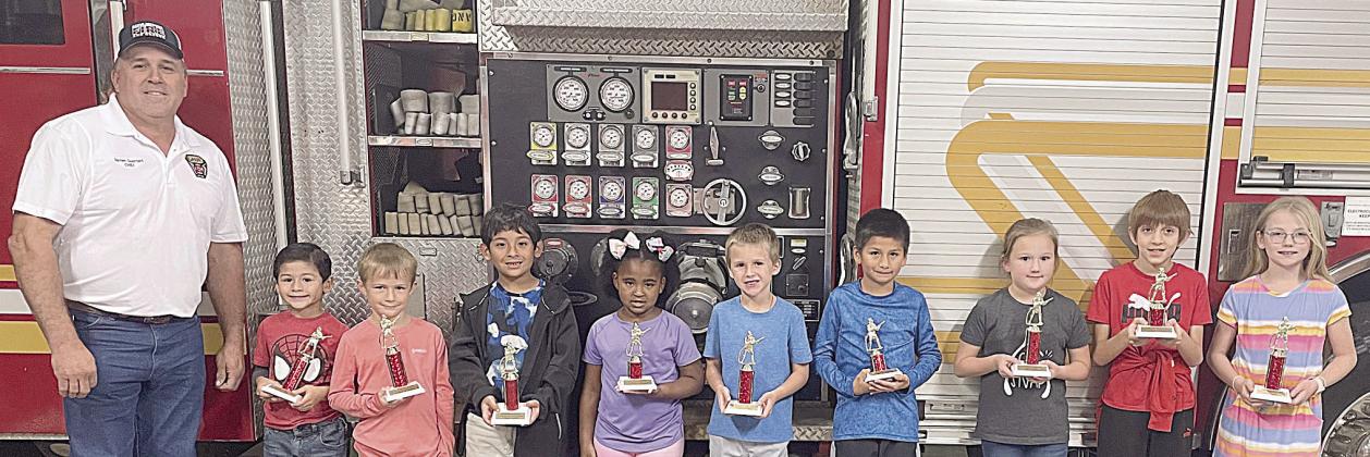 SES WINNERS - From left - Fire Chief Darren Guentert, George Castillo (1st place pre-K), Jenson Kurtz (1st place kindergarten), Eric Ramirez (1st place 1st grade), Paiyton Henry (2nd place 1st grade), Tycen Mendel (1st place 2nd grade), Christopher Castillo (2nd place 2nd grade), JoLee Kuehn (1st place 3rd grade), Greyson Konecny (2nd place 3rd grade), and Selby Harvell (1st place 4th grade).  Not shown – Olivia Otto (1st place 5th grade). Sticker Photo By Darrell Vyvjala