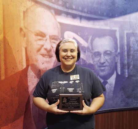 The Edna Christ ian Aca dem y “Best Field Trip” award, which went to the Stanzel Model Aircraft Museum for the 2025-26 school year, is displayed by Cynthia Fogleman, museum director, as she stands in front of likenesses of Victor and Joe Stanzel.