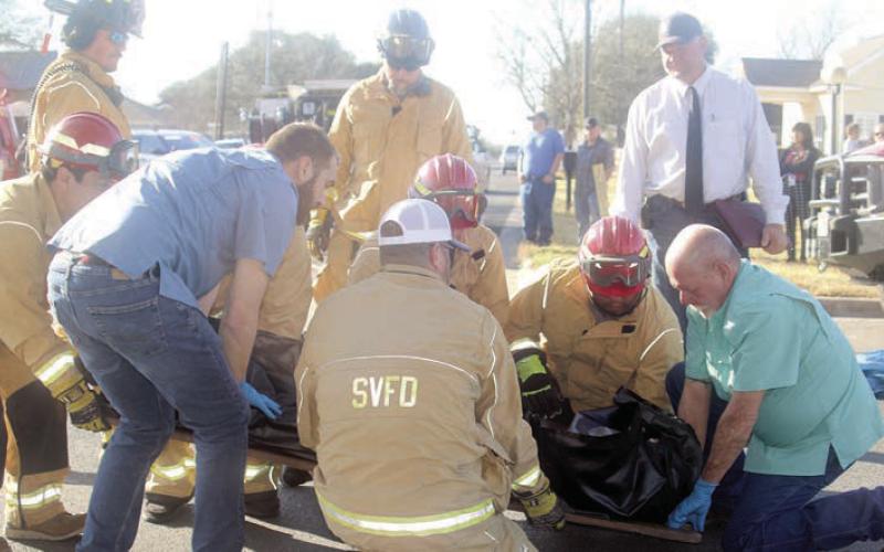 Firefighters assist Clayton Klesel (blue shirt) and Mark Klesel (green shirt) of Schwenke-Baumgarten Funeral Home take away the “deceased” body of Makayla Williams in a body bag. Looking on after conducting an inquest is Justice of the Peace Paul Zapalac (back, right). Sticker Photo By Darrell Vyvjala