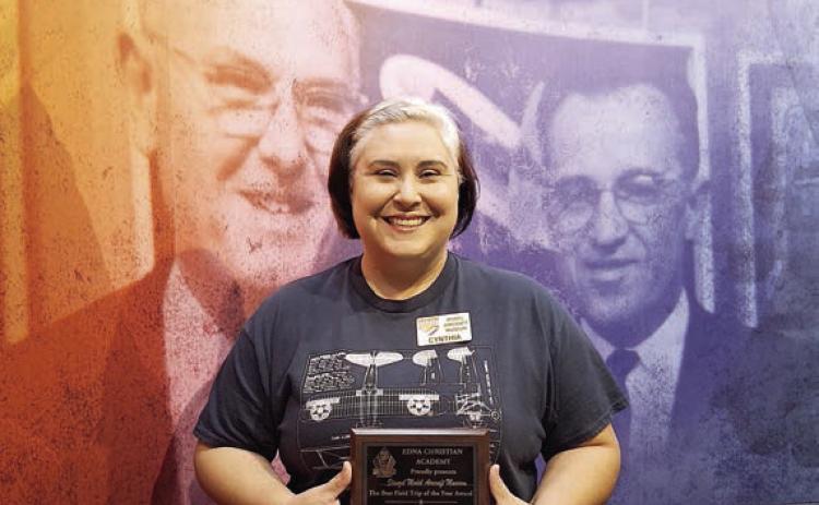 The Edna Christ ian Aca dem y “Best Field Trip” award, which went to the Stanzel Model Aircraft Museum for the 2025-26 school year, is displayed by Cynthia Fogleman, museum director, as she stands in front of likenesses of Victor and Joe Stanzel.
